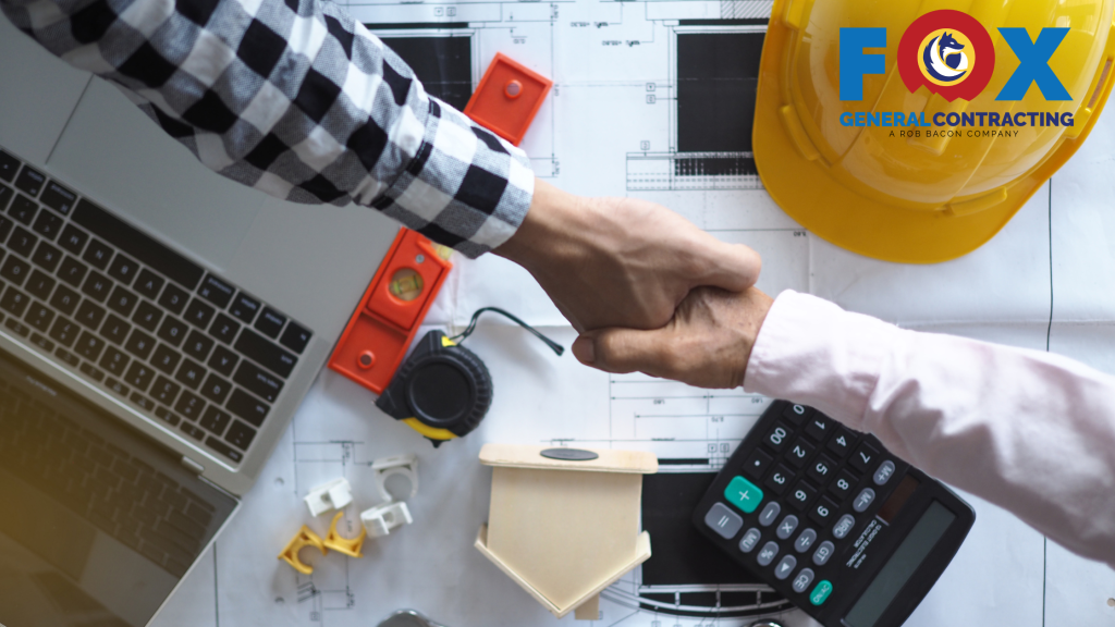 Two people shaking hands over construction plans with tools and a hard hat on the table
